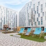 Modern apartment complex with white buildings surrounding a central courtyard. The courtyard features paved walkways, landscaped areas, and a seating area with large blue deck chairs branded "Pavilion Court". Young trees and a wooden gazebo structure are visible in the courtyard space.