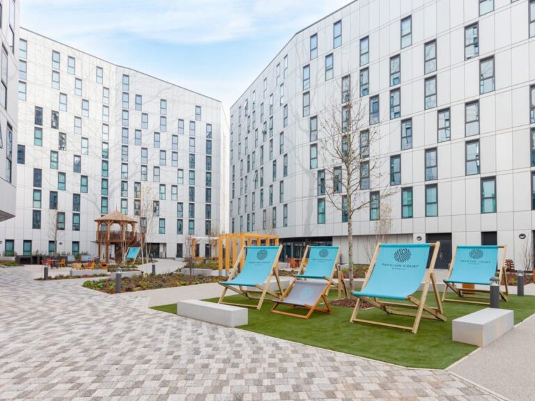 Modern apartment complex with white buildings surrounding a central courtyard. The courtyard features paved walkways, landscaped areas, and a seating area with large blue deck chairs branded "Pavilion Court". Young trees and a wooden gazebo structure are visible in the courtyard space.